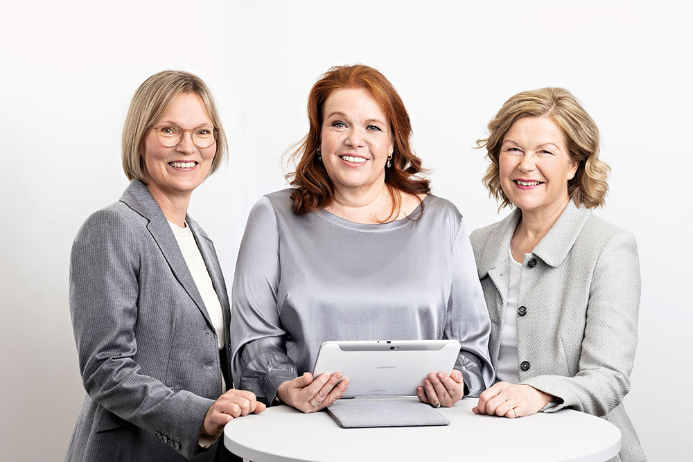 Maarit Tiililä, Sari Ajanko ja Anitta Niemelä seisovat vierekkäin pöydän ja tabletin ääressä tyylikkäissä harmahtavissa vaatteissa.